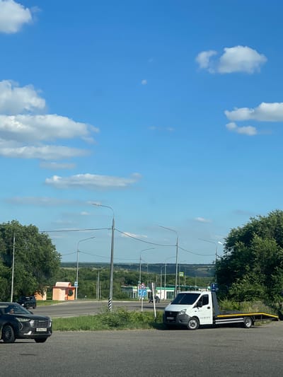 The image depicts a roadside scene with a bright blue sky and scattered white clouds. In the foreground, a black SUV is driving on the road, and a white tow truck with a flatbed is parked on the right side of the road. The tow truck has a license plate that reads "B-967-005-09-99" and a phone number "8-967-005-09-99". In the background, there are green trees, some low buildings, and a road with traffic signs. A small orange bus stop shelter with a curved roof is visible on the left side of the road. Further in the distance, rolling hills are visible under the expansive sky. This scene is identified as being in the city of Donskoe, Russia.
