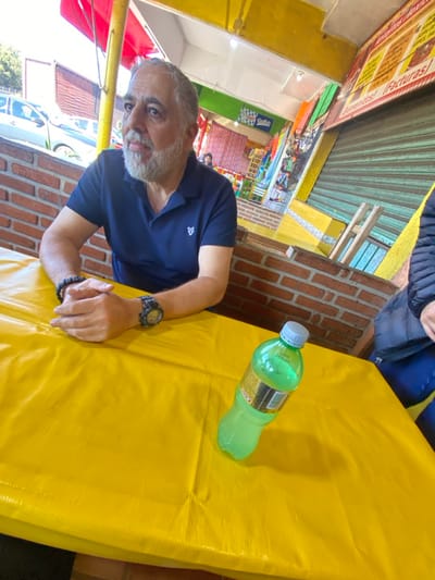 An angled, eye-level outdoor shot shows a man with graying hair and beard, wearing a dark blue collared shirt, seated at a bright yellow table. A green bottle of soda sits on the table. The man wears a watch and beaded bracelet on his wrists, and his hands are clasped on the table. Background includes a store with signage, red and yellow awnings, brick walls, and glimpses of a vehicle outside. Another person is partially visible in the upper right corner.