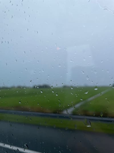 The image presents a blurred, rain-affected view of a landscape in Leidschendam, Netherlands, observed through a window heavily covered with distinct raindrops. The immediate foreground is dominated by the sharply focused water droplets adhering to the glass surface, ranging in size and distribution. Beyond the window, the scene is indistinct due to the rain and blur. The upper portion of the frame shows a muted, overcast sky with a faint, diffuse reddish-orange light source perceptible in the upper-central area. Below the sky, an expansive, blurred green field, indicative of agricultural land, stretches horizontally. A dark, wet roadway or paved surface traverses the lower part of the image, separated from the green fields by a narrow strip of similar vegetation. A single, distinct white line is visible on the roadway, marking a lane or edge. No individuals, explicit actions, or interactions are discernable within the scene.