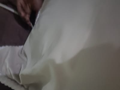 This image shows a close-up, angled view of a light-colored piece of fabric, possibly clothing or bedding, draped over a dark surface. The fabric is creased and has a slightly textured appearance, with subtle folds and shadows creating a sense of depth. The lighting is dim, suggesting an indoor setting, possibly at night. The top edge of the fabric appears to have a decorative trim or seam, with a ruffled or scalloped edge. In the lower right corner, a hand with visible fingers is partially visible, reaching towards the fabric and holding a long, thin object that could be a pen or a stylus. The hand and the object are out of focus. The overall impression is of a casual, intimate moment, possibly related to a craft, writing, or simply adjusting the fabric. The setting in Kipala Mpakani, Tanzania, does not provide specific visual cues within the frame itself, but it informs the broader context of the scene.