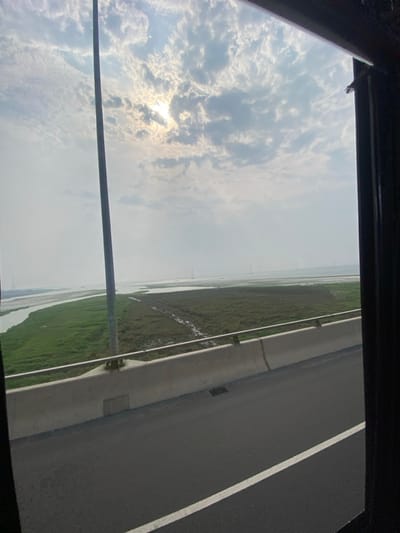 The image displays a view from a vehicle in Naodoba, Bangladesh, looking out through a window. In the immediate foreground, a dark asphalt road with a distinct white lane marker is visible. A light-colored concrete barrier with a metal railing separates the road from the adjacent landscape. A tall, dark, vertical pole, likely a structural component, ascends from the bridge level into the sky on the left-center. Beyond the barrier, a body of water is present on the left, flowing alongside an extensive area of flat, green marshland or low-lying vegetation. Multiple narrow, unpaved channels traverse this vegetated expanse. In the distant background, a large body of water extends towards the horizon, where several faint, slender, dark vertical structures, potentially power transmission towers or bridge supports, are visible. The sky is largely overcast with scattered clouds, and the sun is observed shining through a break in the clouds in the upper left, providing ambient illumination. No people, animals, or active interactions are depicted.