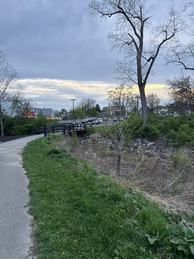 The image depicts a sidewalk in Grove City, Ohio, adjacent to a grassy area and a fence. Beyond the fence, there is a view of buildings and vehicles, suggesting a residential or commercial area. The sky is overcast with some patches of light. Bare trees are visible on both sides of the frame. The overall environment suggests a suburban setting near a park or walkway.