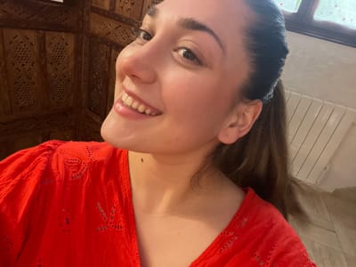 A young adult female is depicted in a close-up, selfie-style photograph taken indoors in Ancenis, France. She has fair skin, dark hair pulled back into a ponytail, and is seen smiling with teeth exposed, looking towards the camera. A small dark mole is present on the lower left side of her neck. She is wearing a bright red top featuring embroidered eyelet details on the sleeves and neckline. The background to her left consists of an intricately carved dark wooden room divider or screen. To her right, a light-colored wall is visible, featuring a white panel radiator and a window with multiple panes, through which blurred greenery can be observed. The scene is illuminated by indoor lighting.