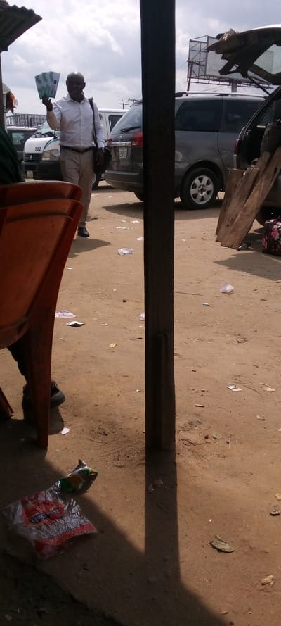 The image shows a scene in Elelenwa, Nigeria, a town with coordinates 4.856 latitude and 7.068 longitude. A man in a white shirt is walking toward the right of the frame. He is holding a stack of green flyers in his left hand and is carrying a brown bag in his right. Behind him are two vehicles. The vehicle closer to the man is a white minivan with a license plate that reads "002." The vehicle further away is a dark grey minivan. The man's feet are on a dirt road that leads away from a large wooden pillar. The wooden pillar casts a shadow on the ground to the right. There is a bright orange plastic chair in the foreground with someone's foot visible in the chair's base. There is a piece of trash on the ground near the bottom of the frame, along with several other pieces of trash visible near the man. There is a sign in the background, but it is too blurry to read. The sky is cloudy. There are power lines in the background.