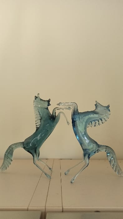 The image displays two translucent, light blue-green glass sculptures, each depicting a rearing horse, positioned on a light-colored surface against a plain, light beige wall. The sculptures are oriented facing each other, with their front hooves elevated and in close proximity, suggesting an interaction. Both figures possess stylized manes and tails. The horizontal surface on which they rest exhibits two distinct vertical seams. The scene is presented indoors, located in the city of Florenville, Belgium.