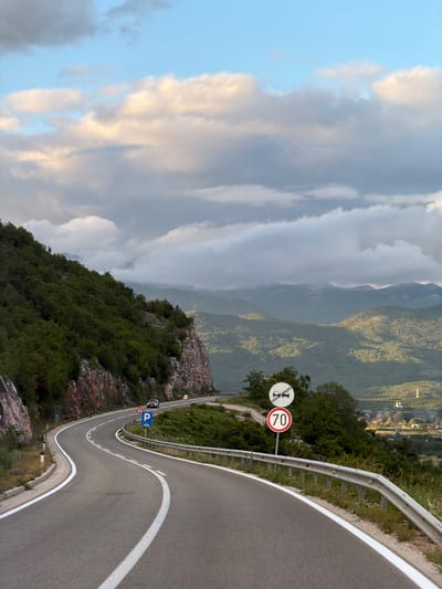 This image captures a breathtaking view of a winding mountain road near Miločani, Montenegro, under a partly cloudy sky during what appears to be late afternoon or early evening. The asphalt road snakes through the mountainous terrain, marked by prominent white lines and protected by a continuous metal guardrail along its right edge.

The landscape is dominated by verdant, tree-covered mountains and deep valleys that recede into a soft haze in the distance. The sky above presents a mix of clear blue and dramatic, textured clouds, some illuminated by the soft sunlight. The immediate roadside is characterized by dense green foliage and rugged rock formations.

Several road signs are visible, guiding travelers. A prominent circular sign indicates a '70' km/h speed limit, alongside another circular sign with a red border prohibiting overtaking (depicting two cars with a diagonal line through them). Further down, a blue rectangular sign displays 'P1', possibly marking a parking spot or viewpoint. A triangular warning sign, likely indicating a sharp curve or other road hazard, is also present.

The scene conveys a sense of serene natural beauty combined with the adventure of navigating a high-altitude mountain pass. The lighting suggests a peaceful transition from day to dusk over this majestic Montenegrin landscape.