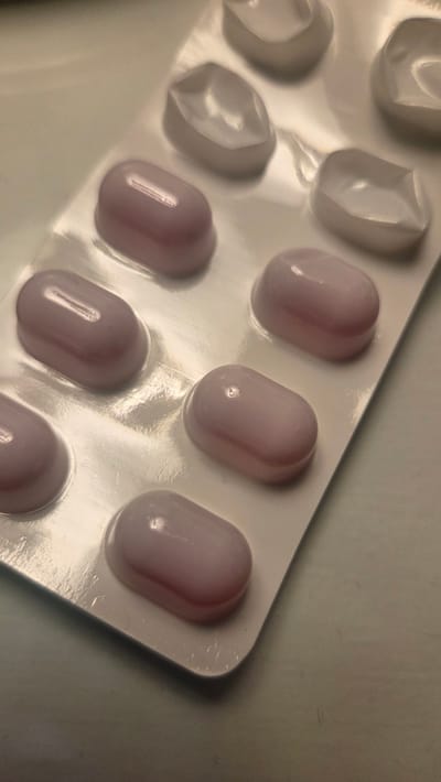 A blister pack of pink oval pills is shown from a slightly elevated angle. The blister pack is made of white plastic with clear plastic coverings over the individual pills. Some of the clear plastic coverings have been removed, revealing empty cavities. The pills themselves are a light pink color and appear to be smooth and coated. The background is a neutral, textured surface, likely a table or countertop. There is no indication of the location in the city of Bray, Ireland.