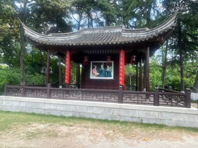 The image depicts a traditional Chinese pavilion in Hailing District, China. The pavilion features a curved, tiled roof, red lanterns, and a low wall with a geometric wooden railing. Inside the pavilion is a framed picture of three individuals. The surrounding environment includes trees and a grassy area.