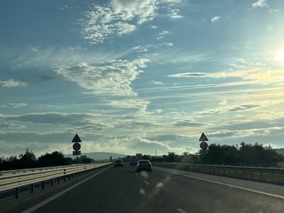 The image shows a view from inside a vehicle driving on a highway. The road is paved with asphalt and has white lane markings. There are guardrails along the side of the road. In the distance, there are trees, hills, and a cloudy sky with the sun shining brightly. There are several cars and a van on the road ahead. Two traffic signs are visible on the left side of the road. One sign is a triangular warning sign with a symbol of a car skidding, indicating a slippery road. Below it is a circular sign with the number "70" in red, indicating a speed limit of 70. Below that is a sign with a snowflake symbol, indicating a warning for icy conditions. The location is identified as the city of Malko Tranovo, Bulgaria.
