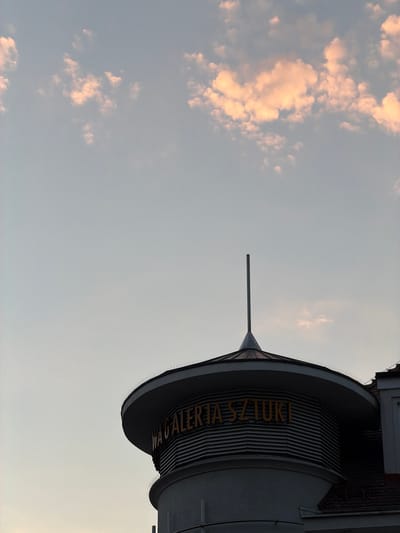 The image depicts the upper section of a building with a curved, layered structure, likely a modern architectural addition to an older building. The building's exterior features horizontal ribbed panels and a conical roof topped with a thin spire. Yellow text is visible on the curved facade, reading "WAGALERIA SZTUKI," which translates to "Art Gallery." Bird spikes are installed along the roofline. The sky above is a pale gradient of gray and blue, with wispy clouds illuminated by a soft, warm light, suggesting either sunrise or sunset. The lower portion of an adjacent building with a tiled roof is visible on the right side. The photograph appears to be taken from a low angle, looking upwards at the building against the sky. The location is identified as the Artus Court (Dwór Artusa) in Sopot, Poland, which houses the Municipal Art Gallery (Miejska Galeria Sztuki).
