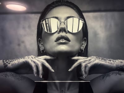 The image is a grayscale, close-up shot of a woman wearing large, reflective sunglasses. Her head is tilted back, and her hands are raised to her jawline, fingers spread. Her arms are covered in intricate tattoos. She has hoop earrings in both ears. Her lips are glossy. A faint, wavy audio visualizer appears at the bottom of the frame. A small cross tattoo is visible on her collarbone. The background is a textured, light-colored wall. There is a circular light fixture in the upper left corner. There is no discernible connection to the city of Krāslava, Latvia.