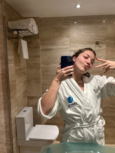 A young woman in a white bathrobe poses for a selfie in a bathroom. She makes a peace sign with her fingers. The bathroom features light-colored tile walls, a towel rack with white towels, and a white rectangular object. The woman holds a blue smartphone to take the photo. The bathrobe has a blue logo on the left side.