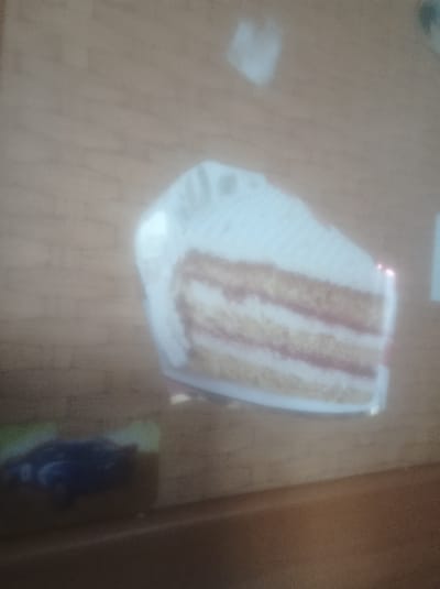 A slice of layered cake with white frosting sits on a wooden surface. The cake appears to have multiple layers of light-colored cake and pinkish filling. A portion of a dark, possibly blue, object resembling a toy car or vehicle is visible in the lower left corner of the frame. The background is a blurred wooden texture, suggesting a table or countertop. The lighting is diffuse, and there are some bright white and faint pink reflections on the surface of the cake. There is no visible text in the image. The overall image is slightly out of focus and has a grainy quality.