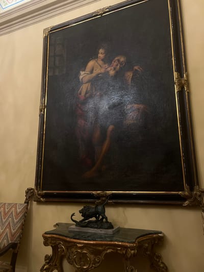The scene is set in an interior room of the Palazzo Negrotto Cambiaso in Sestri Levante, Italy. A large, vertically oriented oil painting is centrally displayed on a light-colored wall. The painting, framed in ornate dark wood with gold leaf accents, depicts the classical subject of "Caritas Romana." It features a young woman with a bare upper torso, draped in fabric, breastfeeding an emaciated, elderly man with a long beard, who is seated and appears weakened. A faint barred window is visible in the dark upper-left background of the painting, suggesting a confined space.
Below the painting, an ornate, gilded console table with a dark green marble top is positioned against the wall. Resting on the marble surface is a dark bronze sculpture mounted on a light-colored rectangular base. The sculpture portrays a lion in an aggressive stance, attacking a coiled snake. To the immediate left, a portion of an antique armchair is visible, featuring a carved wooden frame and upholstery with a multi-colored zigzag pattern. A decorative cornice is present at the intersection of the wall and ceiling in the upper left of the image.