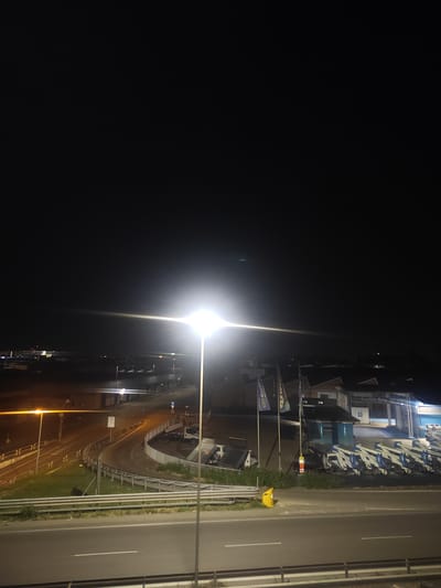 The image captures a nighttime scene in Modena, Italy, likely near an industrial or commercial area given the presence of buildings and a row of what appear to be boats or trailers parked outside. A bright street lamp illuminates the foreground, casting a strong glare. In the midground, a curved road with guardrails is visible, and further back, several buildings with illuminated windows and signage can be seen. The sign on one of the buildings reads "EMILIA GRU". A person wearing a high-visibility vest is standing near a yellow trash bin by the side of the road. The overall impression is of a well-lit area, possibly a business park or a storage facility, on a dark night.