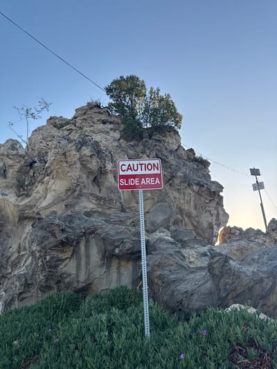The photograph captures a rocky cliffside in Newport Beach, California. A sign reading "CAUTION SLIDE AREA" is prominently displayed. The cliff face exhibits varying shades of gray and beige, with textures indicating erosion and geological stratification. A tree grows atop the cliff. Adjacent to the cliff, vegetation covers the foreground. A cable stretches diagonally across the sky in the background, connecting to another sign affixed to the rock face. The sky is clear and blue.