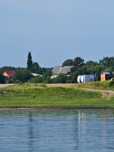 A rural scene unfolds along a wide, calm body of water, likely a river, that occupies the foreground. The water exhibits a blue hue with ripples reflecting the sky and possibly the distant shore. Beyond the water, a grassy embankment rises, dotted with patches of yellow wildflowers and a few dark figures, possibly birds or small animals. A dirt road winds its way across this embankment, separating it from a cluster of buildings and trees in the mid-ground.
The buildings are simple, characteristic of a village setting. To the left, a wooden house with a bright red roof stands prominently. Further to the right, a larger, grey-roofed structure with visible windows suggests a more substantial dwelling. Between these, amidst dense green foliage, are glimpses of other structures, including a white cylindrical tank with blue bands and a small brick outbuilding with a blue roof. A tall, slender pine tree stands out against the sky behind the wooden house. The background is dominated by a dense line of green trees, creating a natural backdrop. The sky above is a clear, pale blue.
The location is identified as Galanino, Russia, suggesting a settlement in the Russian countryside. The scene captures a quiet, natural environment with a few signs of human habitation. There are no visible people in the image. The overall impression is one of rural tranquility under a bright, clear sky.