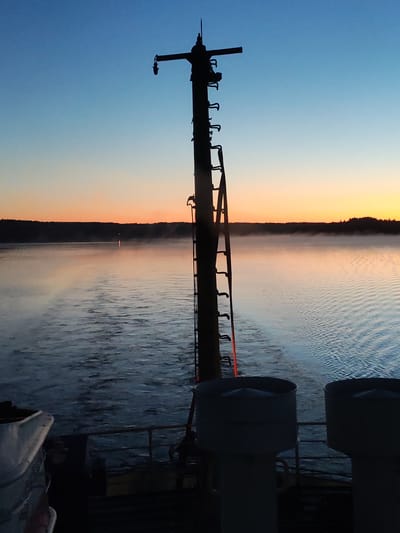 This image captures a pre-dawn or twilight scene from the stern of a vessel on a large body of water, located in the region of Irma, Russia. The sky exhibits a clear gradient, transitioning from a deep twilight blue at the top to a lighter blue mid-sky, and then to vibrant orange and yellow hues closer to the horizon.

A dark, silhouetted landmass, consistent with a forested shoreline, extends horizontally across the distant horizon. The surface of the water in the mid-ground is covered with a layer of atmospheric mist or fog, which appears to reflect the orange light from the sky. A distinct vertical streak of orange-pink light is visible on the water's surface in the distance, likely a reflection of the rising or setting sun or a distant light source. The water immediately behind the vessel shows a clear wake and ripples, indicating the boat is in motion.

In the foreground, a tall, dark, mast-like structure rises centrally from the vessel's deck, casting a prominent silhouette against the sky. This mast is equipped with a vertical series of rungs or hooks resembling a ladder, and near its top, a horizontal arm extends to the left, suspending a dark, downward-facing lantern or light fixture. A faint reddish-orange glow is present on the lower section of the mast and on a dark railing situated below it.

To the bottom left, a section of a white, curved object, possibly a life raft container or similar deck equipment, is visible. To the bottom right, two large, dark, cylindrical structures, appearing to be ventilation shafts or exhaust funnels, rise from the deck. No individuals are visible in the frame.
