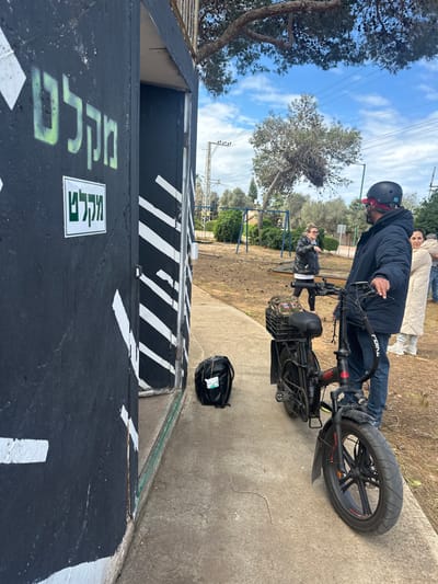 The image displays an outdoor scene in Beit Yitshak Sha'ar Hefer, Israel, featuring four individuals and an electric bicycle on a concrete path next to a building. The building, constructed of dark material and partially visible on the left, features white diagonal stripes and two "מקל"ם" (Miklat - shelter) signs; one stenciled in green and another on a white placard. An open doorway of the building is visible. A black backpack rests on the concrete path near the doorway.
A male subject, wearing a dark puffer jacket, blue jeans, and a black helmet with red accents, stands on the right, holding the handlebars of a black electric bicycle with red accents, fat tires, and a black plastic crate containing a camouflage item mounted on its rear rack. Behind this male, a female subject in a light-colored puffer coat is partially visible, looking towards the left with a slight smile. To her left, a second female subject, wearing a black jacket, white top, and dark pants, stands with her left arm raised in a gesturing motion, facing towards the right. A fourth individual is partially obscured behind the female in the light-colored coat.
The background includes a dirt area with sparse vegetation, a children's playground structure with swings, utility poles with power lines, and several trees under a partly cloudy sky.