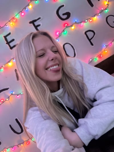A blonde woman in a grey sweatshirt smiles with her tongue out against a white background. Large, dark letters are arranged around her, surrounded by multi-colored string lights. The background resembles the communication wall from "Stranger Things". The image is angled, presenting a slightly tilted perspective.