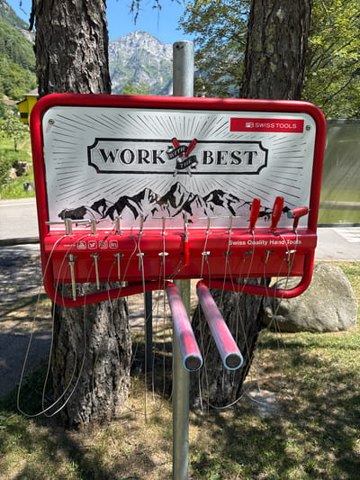 This image captures an outdoor bicycle repair station in Verzasca, Switzerland. The station is mounted on a metal pole positioned between two trees. The central object is a red metal frame holding a white sign that reads "WORK WITH THE BEST PB SWISS TOOLS." Below the sign, a red panel displays various tools suspended by thin wires. These tools include screwdrivers and a tire pump attachment. The background reveals a mountainous landscape, with a road and grassy areas visible. The station appears to be in a park or natural setting, indicated by the trees, rocks, and greenery. There are no people present in the image.