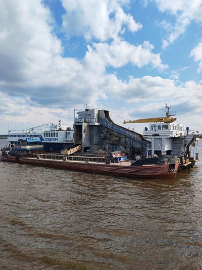Two barges are docked on a murky body of water. The barge in the foreground is primarily rust-colored with white trim. The barge behind it is white and blue, with the word "СЛАВУТИЧ" (SLAVUTICH) emblazoned on its side in blue lettering. This barge also features a large industrial apparatus with a conveyor belt. The scene appears to be situated in a rural or industrial setting, as evidenced by the overcast sky and the presence of these large vessels on the water. The location is identified as Krokhino, Russia.