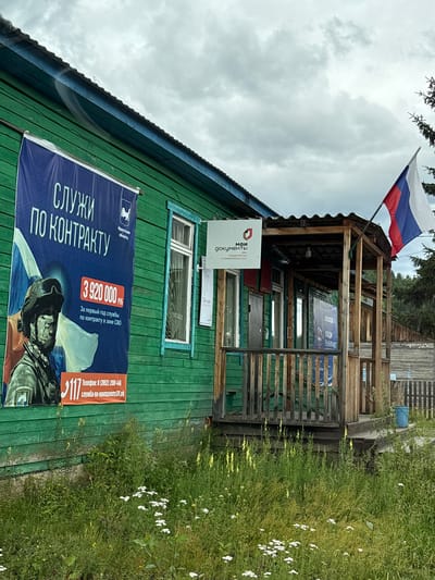 The image shows a green wooden building in the city of Sedanovo, Russia, with a banner on the left advertising military contract service. The banner depicts a soldier in uniform and states that the first year of contract service in the "special military operation zone" (SVO) pays 3,920,000 rubles. Below this, a phone number and website for contract service are listed.
A smaller sign on the building reads "МОИ ДОКУМЕНТЫ" (My Documents), indicating a government service center. The Russian flag is flying from the roof of the building's porch. The building appears to be an older structure, with a wooden porch and railing. Overgrown grass and wildflowers surround the building. The sky is overcast, suggesting cloudy weather. A blue garbage bin is visible on the right side of the image.