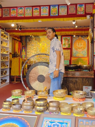The image depicts the interior of a shop in Gokarneshwar, Nepal, filled with Buddhist artifacts. A young man with dark, pulled-back hair stands in the center, wearing a light-colored, short-sleeved shirt with a floral pattern and blue jeans. He is holding a smartphone in his left hand and looking towards the camera.
The shop is decorated with numerous colorful thangka paintings displayed on the walls and above. To the left, a white shelf unit is filled with various sizes of brass singing bowls, some stacked and others placed individually. In the background, a large gong is suspended, with a metal frame around it. Further into the shop, more thangkas, decorative fabrics, and what appear to be prayer flags are visible.
In the foreground, a display table showcases a collection of brass singing bowls of different sizes, each resting on a decorative cushion. The bowls are arranged in multiple rows and appear to be the primary focus of the display. The overall environment suggests a shop specializing in religious and cultural items, likely catering to tourists and devotees. The lighting appears to be artificial, coming from ceiling fixtures.