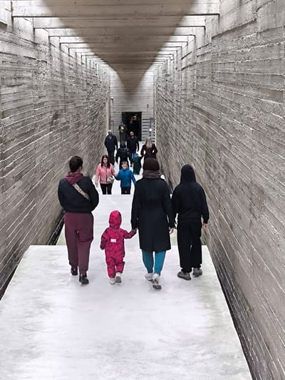 The image shows a group of people walking through the memorial complex in Salaspils, Latvia, which commemorates victims of the Nazi concentration camp. The group is moving away from the camera, down a long, narrow, white-paved corridor defined by tall, rough, concrete walls. The corridor has exposed wooden beams running along the ceiling. A mother and child are in the foreground. Ahead, more people are seen walking in the same direction towards the distant end of the corridor, where a doorway or opening is visible. The lighting is dim, creating shadows and emphasizing the stark texture of the concrete walls.