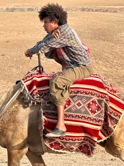 An individual with dark curly hair is seated on a camel, facing left, in the city of Sidi Badhaj, Morocco. The individual is wearing a plaid, long-sleeved shirt, tan trousers, and a gray shoe on their left foot, while the right foot is bare. The camel is covered by a vibrant red and beige patterned blanket. The ground is a sun-baked tan. A metal railing extends from the camel's saddle. The individual is holding onto the railing with both hands.