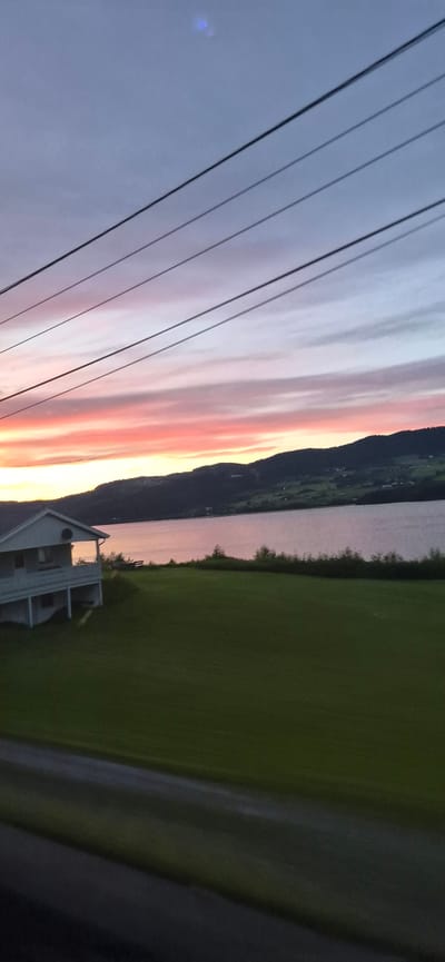 The image depicts a landscape scene in Biri, Norway, during sunset. A vibrant sky painted with hues of pink, orange, and purple streaks across the horizon, transitioning into a soft lavender and grey. In the foreground, a series of dark power lines run diagonally across the frame, partially obscuring the view of the sky.

Beyond the power lines, a body of water, likely a lake or fjord, reflects the colors of the sunset. Along the far shore, a dark, silhouetted mountain range rises, its slopes dotted with subtle lights, suggesting a settlement. To the right of the water, a lush green field stretches out, with a white, two-story house situated near the water's edge. The house has a white railing on its balcony and appears to be situated in a rural or semi-rural setting. The overall atmosphere is serene and tranquil, characteristic of a sunset in a scenic natural environment. The time of day is clearly evening, as indicated by the sunset colors and the soft lighting. No visible text is present in the image.
