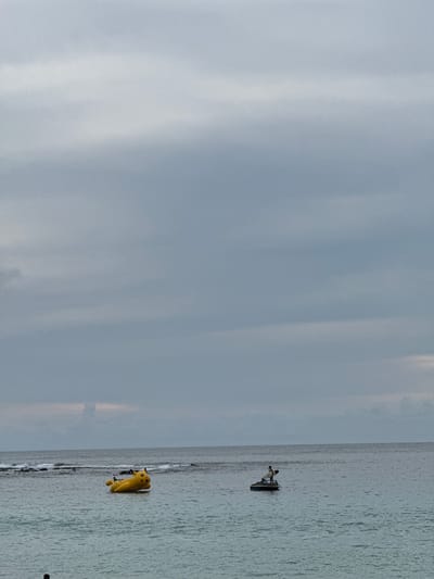 An overcast sky hangs over a body of water where a yellow inflatable Pikachu floats on the left side, and a jet ski with a person holding a surfboard is on the right side. The Pikachu appears to be a large inflatable toy, possibly for recreational use on the water. The jet ski is positioned further out in the water, with a solitary figure standing on it, holding a surfboard. The environment suggests a coastal or marine setting, likely a beach or open sea area, given the presence of waves and the vast expanse of water. The overall mood is calm and somewhat subdued, consistent with the cloudy weather. There are no clear indications of the specific city of Meunasahbale, Indonesia within the image itself, as it is a scene of water and sky with recreational watercraft.