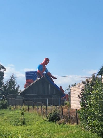 The image shows an inflatable Spider-Man figure perched on top of a wooden structure that resembles a small building with a pitched roof. The Spider-Man figure is red and blue, with its signature mask. The structure appears to be located in a rural or semi-rural area, with a fenced yard in the foreground filled with green grass and some vegetation. In the background, there are trees, a brick wall structure, and a clear blue sky. The overall scene suggests an outdoor recreational area or a residential property with a playful decoration. The image was captured in Блеўчыцкі сельскі Савет, Belarus.