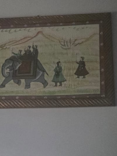 The image shows a framed painting depicting a procession with an elephant carrying riders. The painting is displayed on a wall, with a light-colored wall surface visible above and below the frame. The frame itself is ornate with a woven texture and decorative elements. The painting features a stylized landscape with rolling hills and sparse vegetation. On the elephant, there are at least three figures, one of whom appears to be steering. To the right of the elephant, two figures on foot are depicted, one in a green robe and the other in a dark robe holding a staff. The overall style of the painting suggests it is a historical or cultural representation. The location is not identifiable from the image itself, and therefore, it is not possible to determine its presence in Ladispoli, Italy.
