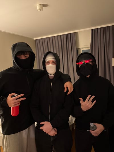 Three individuals are posing indoors. They are wearing black hooded sweatshirts and face coverings. The person at the top has on red flame-shaped sunglasses. The middle person's face is wrapped in bandages. The person at the bottom has a black balaclava. All three are holding objects. The background includes a light-colored wall, a smoke detector, and gray curtains.