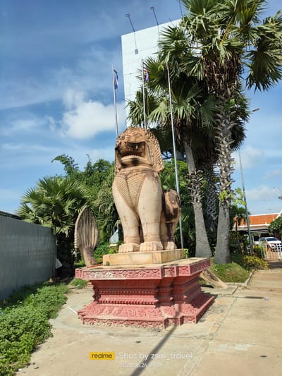 This is a photograph of a stone lion statue in Bavet Quarter, Cambodia. The statue is a prominent subject, positioned centrally on a decorative red pedestal. The lion is carved with intricate details, including a patterned mane and armor-like detailing on its chest and legs. To the left of the lion, a section of a Naga serpent sculpture is visible.
Behind the statue, several palm trees dominate the background, their fronds spread wide against a bright blue sky with scattered white clouds. A tall, white building stands to the left of the palm trees, and flags on poles are visible in front of it. To the right, a building with a red tile roof and a white structure suggest a commercial or public area. A white car is parked on the right side of the image.
The scene is bathed in sunlight, indicating it is daytime, likely late morning or early afternoon given the shadows. The weather appears clear and warm. The ground in the foreground is paved and shows some wear. Green foliage and bushes are present around the base of the statue and along the left side of the image.
The overall impression is one of a historical or cultural landmark situated in a tropical environment. The inscription at the bottom right of the image reads "realme Shot by zsm_travel 2026.06.24 09:19".