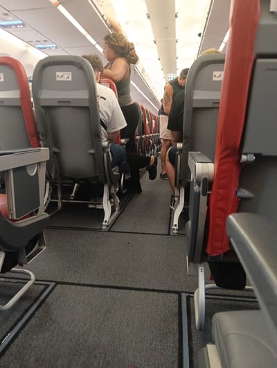 The image displays the interior of an airplane cabin, viewed from an aisle seat looking towards the front or middle of the aircraft. The setting is brightly lit by artificial overhead lighting, illuminating the white ceiling panels and overhead compartments, and the grey carpeted aisle.

Several passengers are visible. In the mid-ground on the left, a woman with curly brown hair, wearing a green tank top and dark pants, stands in the aisle, reaching up towards an overhead compartment, suggesting she is stowing or retrieving luggage. Directly across from her, a man in a dark t-shirt and light-colored shorts, with a tattoo on his right arm, is seen from behind, leaning forward in the aisle. Further down the aisle, more passengers are partially visible.

In the foreground on the left, the back of a grey airplane seat is prominent, with an individual in a white t-shirt and blue jeans seated, seen from behind. A sticker on this seat back clearly displays symbols for "fasten seatbelt" and "upright seat back," indicating safety instructions. Similar instructional symbols are partially visible on a seat further down the right aisle. The overall activity suggests passengers are either boarding or disembarking the aircraft. No specific time of day or weather is discernible from the interior view. The location is an airplane, with the external context suggesting it is in San Bartolomé, Spain.