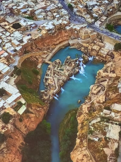 This is a high-angle, aerial-view photograph of an urban landscape with a river or body of water running through it. The water is a vibrant turquoise color and features several sections of white rapids, indicating a dam or waterfall.
The surrounding environment is a mix of natural and man-made structures. On the left side of the image, there is a densely populated urban area with many buildings. These buildings have light-colored walls and flat roofs, arranged in a somewhat irregular pattern. A road or pathway with parallel lines runs through this urban area, suggesting transportation infrastructure.
The center of the image is dominated by the water and several structures built within or around it. These appear to be ruins or ancient buildings, with a complex layout of walls and courtyards. The architecture suggests historical significance, possibly an old city or settlement.
To the right of the water, the landscape transitions to rugged, rocky terrain with patches of green vegetation, suggesting a more natural and uncultivated area. Further to the right, more buildings are visible, some with distinct white roofs.
The lighting suggests it is daytime, with shadows indicating the sun is not directly overhead. The overall scene is one of a historical settlement integrated with a natural water feature. There is no visible text in the image.