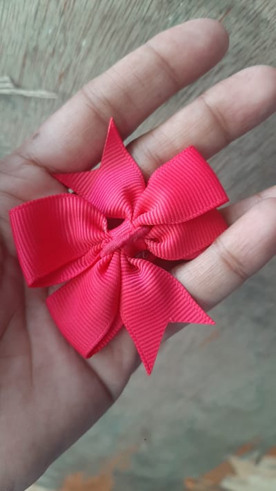 A close-up, overhead shot shows a bright pink grosgrain ribbon bow held in a person's open palm against a wooden surface. The bow is intricately folded with multiple loops and pointed ends, creating a star-like shape. The person's skin is visible on the palm and fingers, with clear fingerprints and texture. The background is a wooden surface, likely a table or floor, with a visible wood grain pattern. The lighting suggests an indoor setting with soft, diffused light. No specific location within Kawit, Philippines is discernible from the image itself.