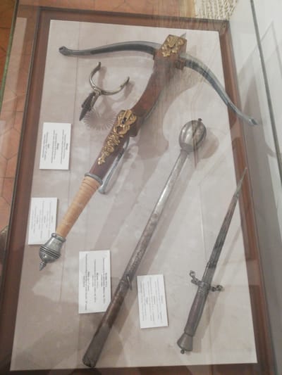 The image presents a top-down view of historical weaponry displayed in a rectangular glass museum case with a dark brown wooden frame, situated within the Mir Castle Complex Museum in Мірскі сельскі Савет, Belarus. The interior of the display case features a light-colored, off-white or beige background surface.
Four primary objects are arranged within the case. Dominantly positioned diagonally from the upper left to the lower center is a crossbow. It features a wooden stock with ornate gold-colored metal plating, a dark metal bow (prod), and a light-colored, possibly leather-wrapped, grip on the rear section. To the upper left of the crossbow's stock, a smaller, intricate silver-colored metal object, resembling a spur or a crossbow cocking aid, is present.
Below the crossbow, a long, slender, silver-colored metal mace with a distinct fluted, spherical head is laid out diagonally. To the lower right, a shorter, pointed metal dagger or stiletto, complete with a hilt and guard, is displayed. Four rectangular white informational placards, bearing text in Belarusian, are distributed strategically within the case, adjacent to the artifacts. Reflections from overhead lighting are visible on the glass surface of the display case. No individuals are depicted, and the objects are static exhibits.