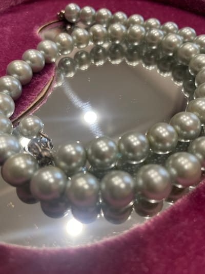The image shows a close-up, high-angle shot of a pearl necklace with a vintage clasp. The necklace is resting on a pink, velvety surface. The pearls are round and appear to be a pale gray or silver color, with a lustrous sheen. The clasp is ornate and metallic, with intricate detailing. The background is a reflective surface, likely a mirror, which shows the pearls and the pink velvet surface from different angles. There are bright light reflections on the mirror, suggesting artificial lighting. The overall aesthetic is one of elegance and sophistication, with a vintage feel. No people or specific location cues are visible, beyond the general context provided of Aropakkuzi, Russia. The time of day, weather, and emotions are not discernible from the image. There is no visible text in the image.