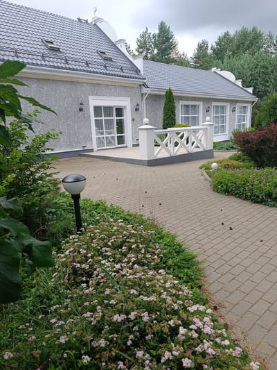 The image depicts a modern gray house with a dark gray tiled roof, featuring multiple windows with white trim. A paved walkway with interlocking gray pavers leads around the house, bordered by green vegetation and flowering shrubs. A black lamppost with a white spherical shade stands near the walkway. A white decorative railing with a lattice design is situated on a raised platform. The background consists of tall evergreen trees, suggesting a semi-rural or suburban environment. The scene is outdoors, with a cloudy sky overhead. The location is identified as Starobin Village Council, Belarus. There are no people or animals visible in the image.