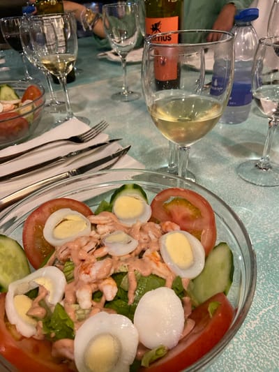 The image shows a table setting in a restaurant located at 28.738, -13.867 in Corralejo, Spain. A glass of white wine is partially filled, sitting next to a glass of water and a bottle of white wine. The table has a light blue tablecloth. A glass bowl filled with salad is in the foreground. The salad contains shrimp, lettuce, tomato, cucumber, and hard-boiled eggs. The salad is dressed in a creamy pink sauce. There are two forks and a knife on the table. A white napkin is partially visible behind the salad bowl. The table is partially visible in the background.