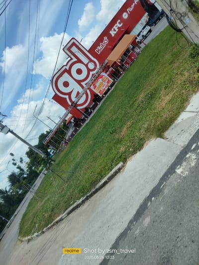 The image shows a street view of a KFC restaurant and a large "Go!" sign in Gò Dầu, Vietnam. The KFC restaurant building is red with white lettering. A white SUV is parked in front of the KFC. The "Go!" sign is large and red and white, with a stylized "Go!" logo. The street is paved and lined with grass. There are utility poles and wires in the background. The sky is blue with white clouds. The image is taken from a slightly tilted angle.