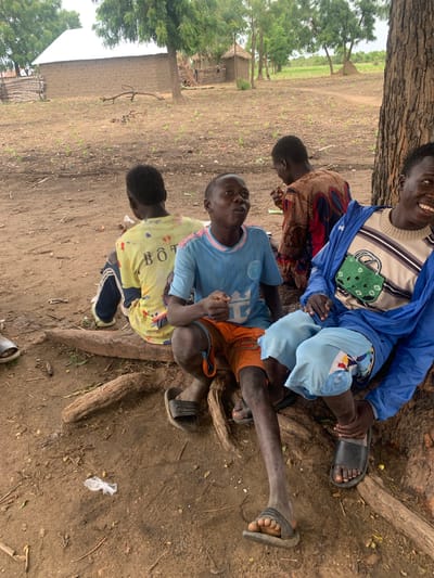 Four young boys are gathered near the base of a large tree in an outdoor area. The ground is dirt with scattered dry leaves and twigs. In the background, there are simple structures made of mud and thatch, as well as some greenery and trees. The boys are casually dressed. One boy, in the foreground to the right, is wearing a blue jacket over a striped shirt and light blue shorts, and he is smiling and looking to the side. The boy next to him, slightly to the left, is wearing a light blue t-shirt and orange shorts, and he is looking forward with his mouth slightly open. Another boy, seated further back and to the left, is wearing a yellow t-shirt with writing on the back and dark pants. The fourth boy, in the middle background, is wearing a patterned brown and red garment and is holding something to his mouth. The scene appears to be in a rural setting in Dull A, Nigeria.