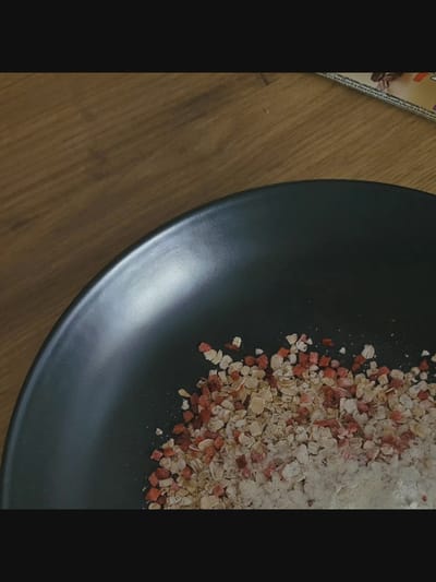 The image shows a close-up view of a dark, matte ceramic bowl containing a mixture of what appears to be rolled oats and small, freeze-dried red fruit pieces, likely strawberries, as well as some powdered substance. The bowl is placed on a wooden surface with a visible grain pattern. The lighting suggests an indoor setting. The location is noted as Bielawska Wieś, Poland, though no specific landmarks or contextual elements within the image confirm this. There are no people or animals visible in the image, and no actions or interactions are taking place. The overall scene is static and focuses on the food ingredients in the bowl.