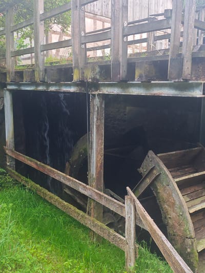 The image shows a close-up of an old water mill structure. In the foreground, there is green grass and moss-covered wooden railings. Behind the railings, a large wooden water wheel is partially visible within a dark opening, with water flowing over it. The structure above the water wheel is made of weathered wood and metal beams. The background reveals part of a building with timber framing and wooden planks. The overall impression is of an aged, rustic, and somewhat derelict industrial facility. The scene is located in Lügde, Germany, specifically at the historic "Papiermühle" (Paper Mill).