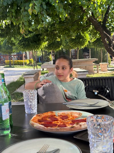 A young girl is seated at a table outdoors, likely at a restaurant or cafe given the presence of food and drinks. She is wearing a light green t-shirt with a white flower graphic. The girl is looking towards the camera with a gentle expression. On the table in front of her are plates with cutlery, a pizza with pepperoni, a crystal glass, and two green glass bottles of water. One bottle's label is partially visible, showing "BY" and "REAL WATER" in text.

The scene is set outdoors, with abundant green foliage and trees in the background, suggesting a sunny day with pleasant weather. The setting appears to be a garden or a terrace area, with stone pathways and benches visible. The time of day appears to be daytime, given the lighting. The overall mood is relaxed and casual, indicative of a meal shared with others, although only the girl is fully visible. The location context is Ashtarak, Armenia.