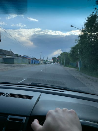 The image depicts a view from the driver's seat of a car looking forward through the windshield. A person's right hand is visible on the steering wheel. The car is on a paved road with a white lane marking. To the left of the road, there are houses with corrugated metal roofs and fences, and utility poles with wires. To the right, there are trees and more houses. In the distance, traffic lights are visible, showing red. The sky is cloudy with sunlight breaking through in patches. The scene appears to be in a residential area of a city.