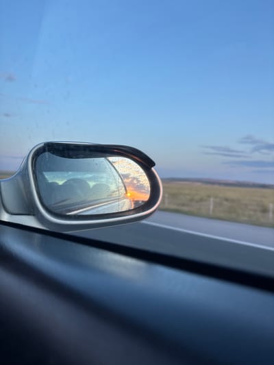 The image captures a view from inside a vehicle, looking into the side-view mirror, which reflects a sunset. The mirror is slightly fogged with condensation, obscuring some details of the reflection. The reflection shows a bright orange sun low on the horizon, partially obscured by clouds, casting a warm glow across the sky. In the foreground of the mirror's reflection, several indistinct, rounded shapes, possibly passengers or car interior elements, are visible. Outside the mirror, the landscape is a blur of green fields and a hazy sky. A road is visible in the distance, with fence posts lining its edge. The vehicle's interior, particularly the dashboard and door panel, is visible in the lower portion of the image, appearing dark and reflective. The overall scene suggests travel during a sunset. The location is identified as Palauzovo, Bulgaria.